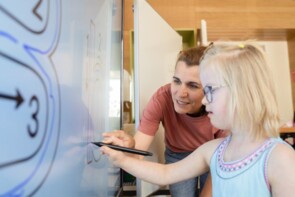 Eine junge Schülerin steht vor einer digitalen Tafel und zeichnet darauf mit einem Stift. Eine Frau lehnt sich zur ihr hinunter und unterstützt sie beim Zeichnen.