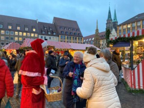 Marcel Schneider mit rotem Umhang und roter Nikolausmütze verkleidet unterhält sich mit drei älteren Damen. Eine davon strahlt den Friseurmeister an. Im Hintergrund sieht man die Nachbarstände.