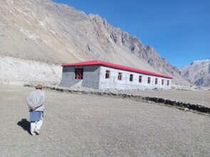 Das neue Schulhaus steht auf einem Hochplateau mitten im Nirgendwo, vor einem hohen Gebirgszug. Vorne im Bild läuft ein einzelner Mann auf das grau-verputzte Gebäude mit rotem Dach zu.