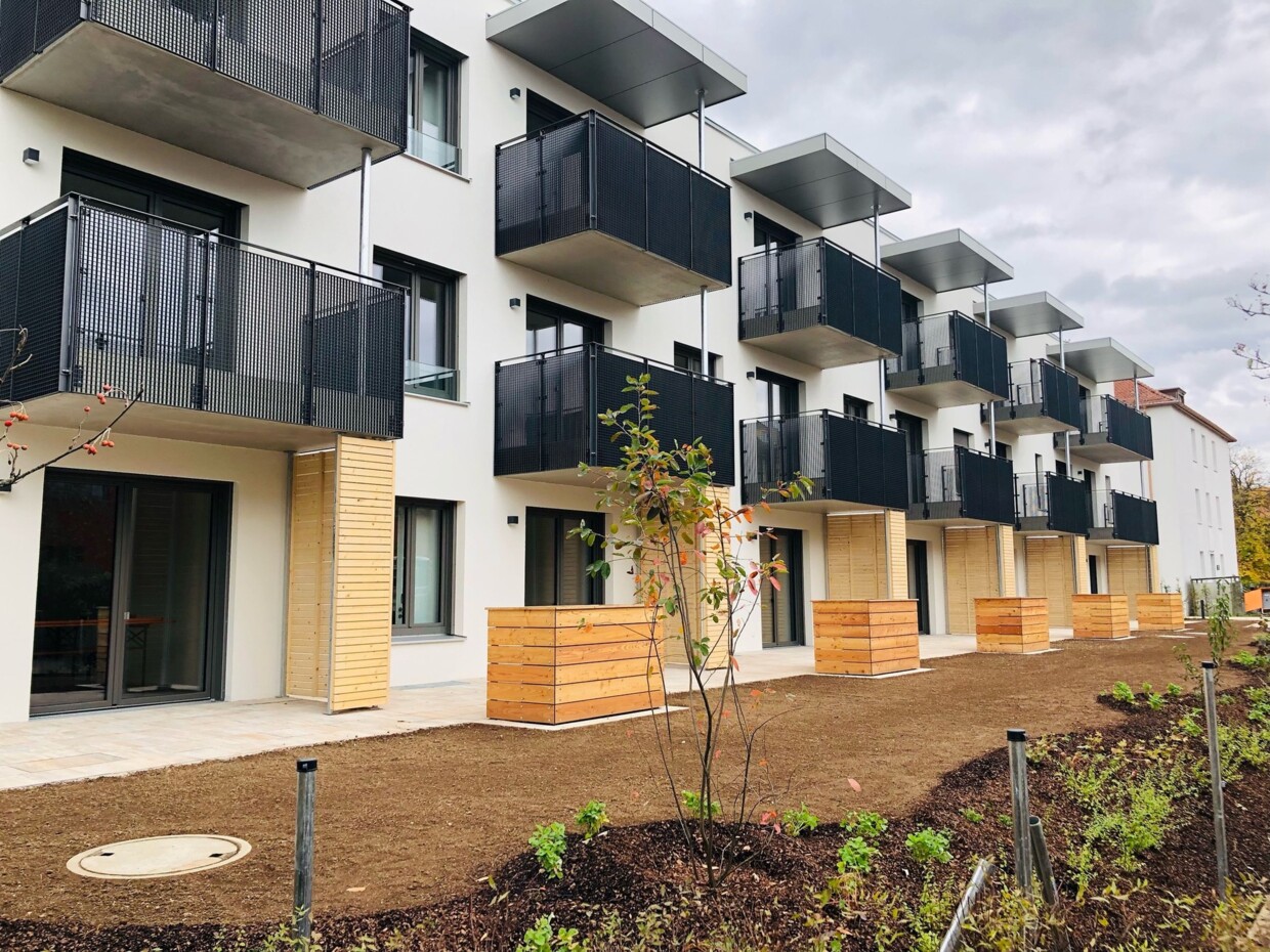 Die Rückseite der Appartementanlage besteht aus einer hellen Fasse, Holzelementen und Balkonen mit einer schwarzen Metallbrüstung. Man sieht außerdem den frischangelegten Garten beziehungsweise den Rasenbereich und die Hochbeete.
