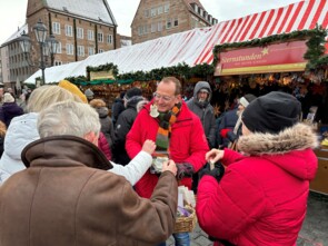 Volker Heißmann umringt von einer Traube älterer Marktbesucherinnen und Marktbesucher verteilt Sterne und Engel gegen Spende.