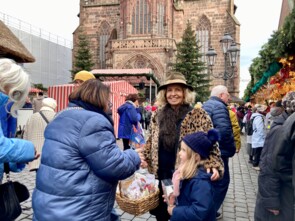 Eine Markbesucherin sucht sich aus dem Körbchen von Michaela May einen Stern aus. Michaela May schaut in die Kamera und lächelt. Im Hintergrund sieht man die Nürnberger Frauenkirche.