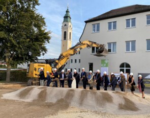 Eine Gruppe von dreizehn Menschen mit Helmen auf dem Kopf steht hinter einem flachen Haufen aus Kies und Sand. Sie halten Spaten in den Händen und führen den Spatenstich durch. Im Hintergrund stehen ein Banner der KJF und ein gelber Bagger.