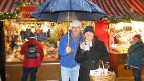Karin Rabhansl steht mit gefüllter Spendendose vor dem Sternstunden-Stand. Weil es regnet, hält ihr Kilian Greger von der Versicherungskammer einen Schirm. Beide lächeln in die Kamera.