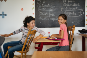 Zwei Mädchen sitzen an ihren Schulbänken. Die Linke kippelt auf ihrem Stuhl und dreht sich zu ihrer Mitschülerin nach hinten um, diese blickt in die Kamera. Beide lachen.