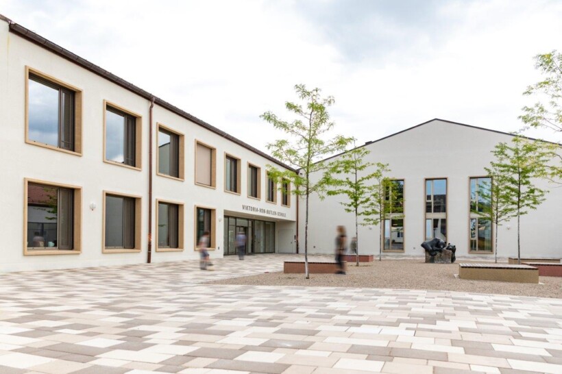 Blick auf den Hof und den Eingangsbereich der neuen Schule. Die Fassade ist hell und der Hof ist mit Platten in Weiß- und Brauntönen gepflastert. Rechts im Bild sieht man 4 frisch gepflanzte Bäume.