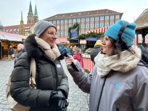 Marianne Lüddeckens von Sternstunden gibt der Bayern 1-Reporterin ein Live-Interview. Im Hintergrund sieht man Nachbarbuden auf dem Christkindlesmarkt und das Nürnberger Rathaus.