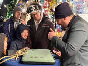 Der Zauberer steht vor einem Bistrotische auf dem seine Zauberutensilien bereit legen. Er zeigt gerade einer Familie mit zwei Kindern einen Zaubertrick.