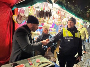 Der Zauberer links im Bild hält dem Polizisten rechts im Bild eine Spielkarte hin. Der Polizist versucht sie an der richtigen Stelle zu greifen. Im Hintergrund steht eine Frau von Sternstunden mit einer Spendendose.