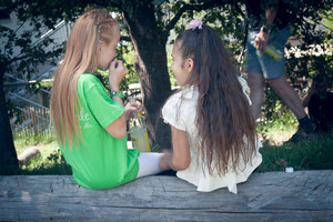 Zwei Mädchen sitzen mit dem Rücken zur Kamera auf einem Baumstamm, das Mädchen links riecht an einem grünen Papier und hat eine Limoflasche in der Hand, das Mädchen rechts sieht dabei zu