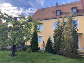 Ein oranges-gelbes Haus versteckt sich hinter einem Obst- und einigen Nadelbäumen. Es ist das Haus, das umgebaut werden soll.