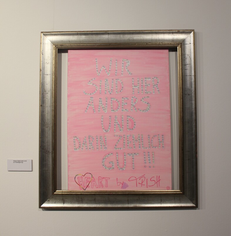 Das Motto der Ausstellung "Wir sind hier anders und darin ziemlich gut!" auf rosa Leinwand mit silbernem Rahmen.