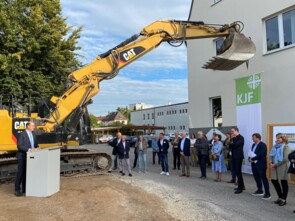 Links im Bild steht der Staatsminister Albert Füracker an einem Rednerpult auf dem Gelände des Neubaus und hält eine Rede. Rechts steht das Publikum und hört aufmerksam zu. Im Hintergrund steht ein großer gelber Bagger.