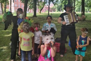 Eine Veranstaltung im Park: Kinder tragen selbstgebastelte Feder-Masken. Zwei Frauen betreuen die Kinder, eine spielt Akkordeon.