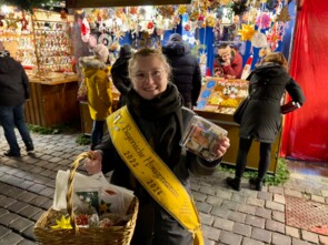 Linda Jakob trägt eine goldene Krone und eine gelbe Schärpe mit der Aufschrift "Bayerische Honigprinzessin 2022-2024". Sie hält stolz ihre volle Spendendose in die Kamera.