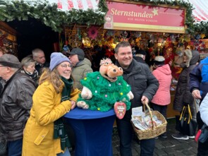 Eine Besucherin macht ein Foto mit Sebastian und Amanda, die auf einem blau-umhüllten Bistrotisch sitzt. Im Hintergrund sieht man den Sternstunden-Stand.