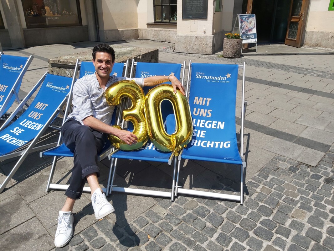 Dominik Glöbl sitzt auf einem der Sternstunden-Liegestühle, neben ihm zwei weitere Liegestühle und Heliumballons, die eine 30 zum 30-jährigen Jubiläum bilden. 