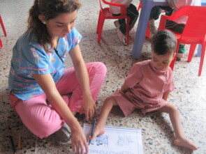 Eine Betreuerin der Einrichtung unterstützt einen kleinen Jungen, der einen Stift zwischen seinen Zehen hält und malt, weil er seine Arme auf Grund einer Behinderung nicht nutzen kann.
