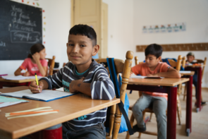 Schülerinnen und Schüler sitzen an ihren Schulbänken und schreiben in ihre Hefte. Der Junge in der vordersten Reihe blickt direkt in die Kamera. 