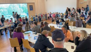 Im Vordergrund sitzen Besucherinnen und Besucher an Tischen, vor ihnen Getränke. Im Hintergrund sieht man die Kinder des Kindergartens Kicherkiste mit bunten Capys auf den Köpfen bei ihrem Auftritt.