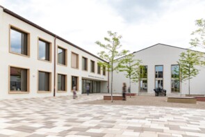 Blick auf den Hof und den Eingangsbereich der neuen Schule. Die Fassade ist hell und der Hof ist mit Platten in Weiß- und Brauntönen gepflastert. Rechts im Bild sieht man vier frisch gepflanzte Bäume.