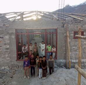 Eine Gruppe von Kindern steht vor den Mauern ihrer neuen Schule. In diesem Foto fehlt noch das Dach und der Verputz. Im Hintergrund sind hohe Berge zu sehen.