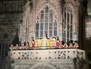 Das neue Nürnberger Christkind Nelli Lunkenheimer steht auf dem Balkon der Frauenkirche am Hauptmarkt und eröffnet mit geöffneten Armen und einem großen Lächeln den Christkindlesmarkt. Umrahmt wird das Christkind von Blasmusikern und zwei kleineren Engeln jeweils links und rechts.