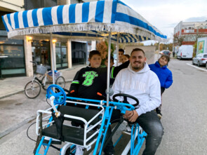 Das Mehr-Personen-Tandem der Sozialeinrichtung hat ein blau-weiß-gestreiftes Dach. Es sitzen zwei Jungen vorne und vier weitere Kinder auf der Rückbank.