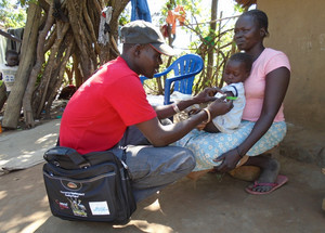 Mann (links) misst Oberarmumfang bei einem Kleinkind (Kontrolle des Ernährungszustands), Kind sitzt bei Mutter auf dem Schoß 