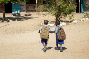 Zwei Mädchen (von hiten) mit großen braunen Rucksäcken gehen nebeneinander zum Schulgebäude