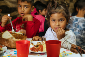 Zwei kleinere Kinder, ein Junge und ein Mädchen, essen ein warmes Essen. Sie blicken in die Kamera.