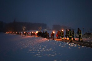 Auf einem Weg im Schnee mit Wald im Hintergrund sind man eine große Gruppe von Menschen mit Fackeln den Hügel hinauswandern.