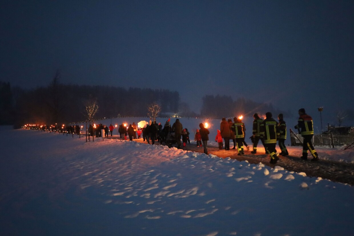 Auf einem Weg im Schnee mit Wald im Hintergrund sind man eine große Gruppe von Menschen mit Fackeln den Hügel hinauswandern.